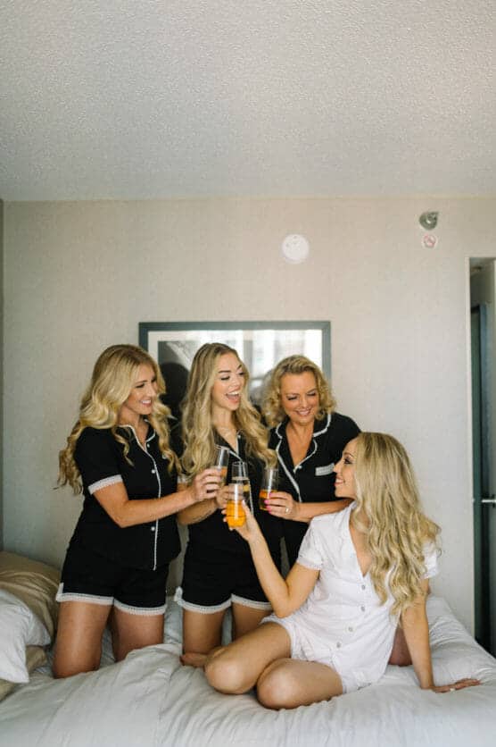 Four women in matching pajamas smile and toast with drinks while sitting and kneeling on a bed in a bright, modern room. Three wear black pajamas, and one wears white. They appear to be celebrating together. by Justin Salem Meyer