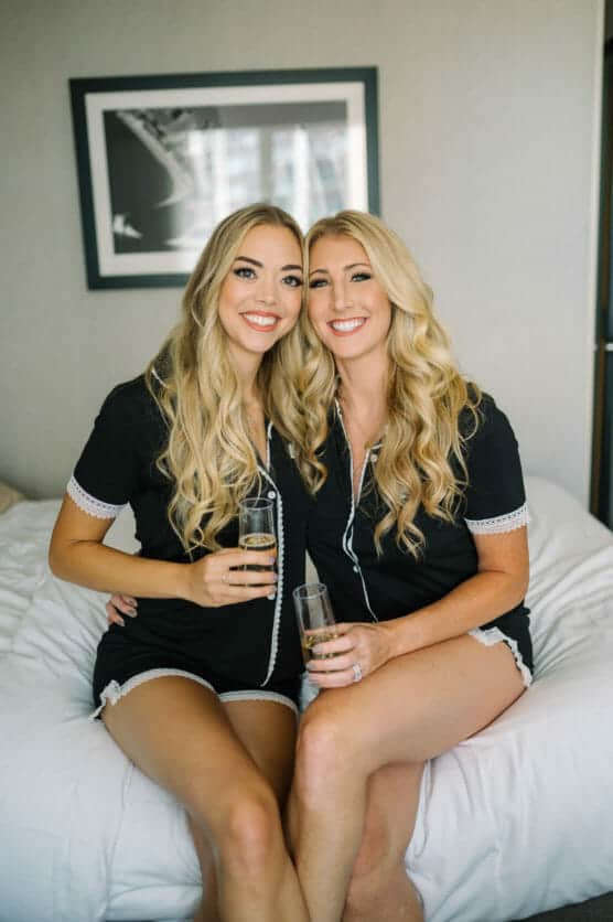 Two women with long blonde hair, wearing matching black pajama sets, sit closely together on a bed, smiling and holding glasses of champagne in a bright, modern room. by Justin Salem Meyer