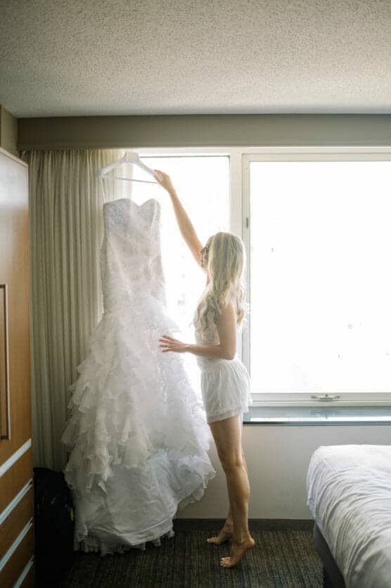 A woman with long blonde hair stands in a sunlit room, reaching up to hold a white, ruffled wedding dress hanging near a window. She is wearing light-colored sleepwear and barefoot on the carpeted floor. by Justin Salem Meyer