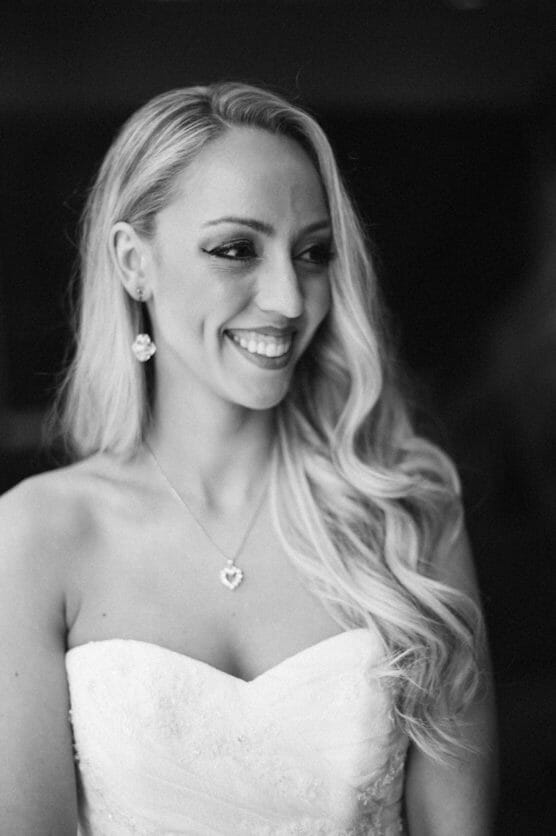 A black-and-white photo of a smiling woman with long, wavy blonde hair in a strapless dress, wearing heart-shaped earrings and a matching necklace. She looks to the side with a joyful expression. by Justin Salem Meyer