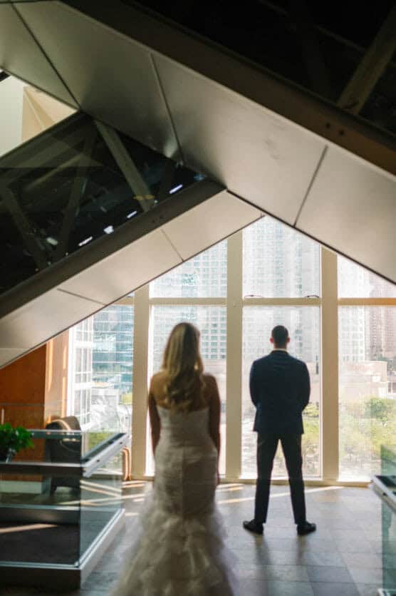 A bride in a white dress stands indoors, looking at a groom in a suit who faces large windows with a cityscape view, both framed by modern architectural elements. by Justin Salem Meyer