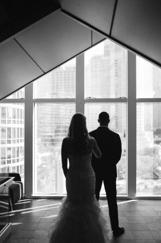 A bride and groom, seen from behind in silhouette, stand side by side facing large windows with a cityscape of tall buildings visible outside. The room is modern with geometric ceiling lines. by Justin Salem Meyer