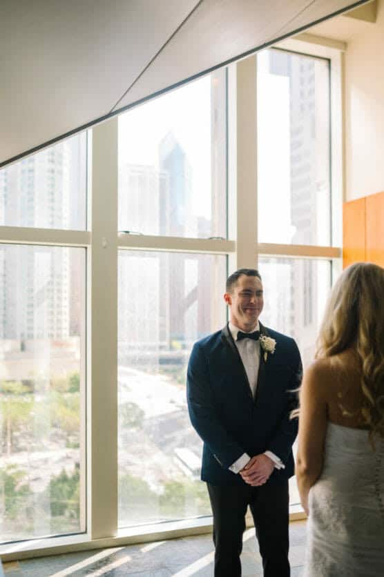 A groom in a dark suit and bow tie smiles at a bride with long blonde hair. They stand indoors by large windows with a city skyline visible outside on a sunny day. by Justin Salem Meyer