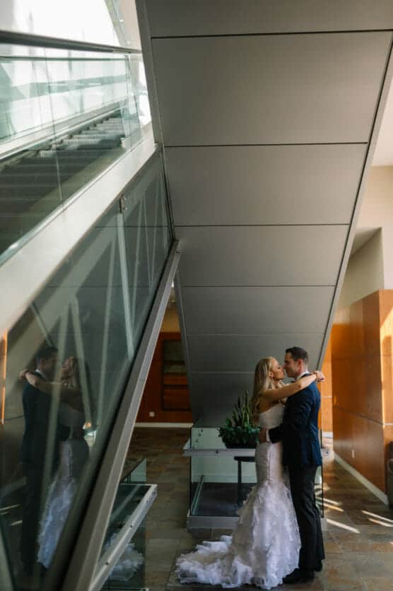 A bride and groom embrace under a modern, angular staircase with their reflection visible in the glass beside them. Natural light fills the contemporary indoor setting. by Justin Salem Meyer