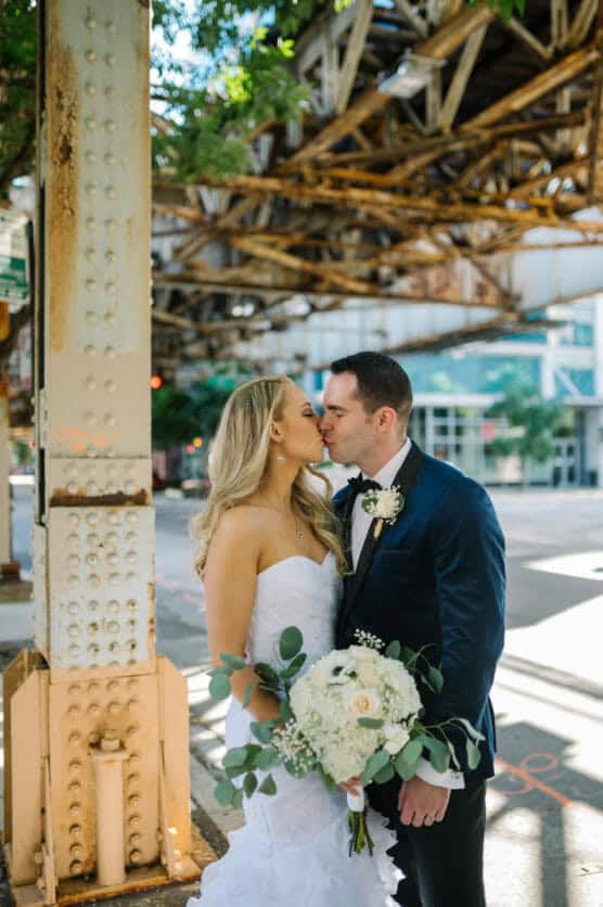 A bride and groom share a kiss beneath an urban elevated train track. The bride holds a white bouquet and wears a strapless dress, while the groom wears a dark suit with a boutonniere. Sunlight filters through the structure. by Justin Salem Meyer