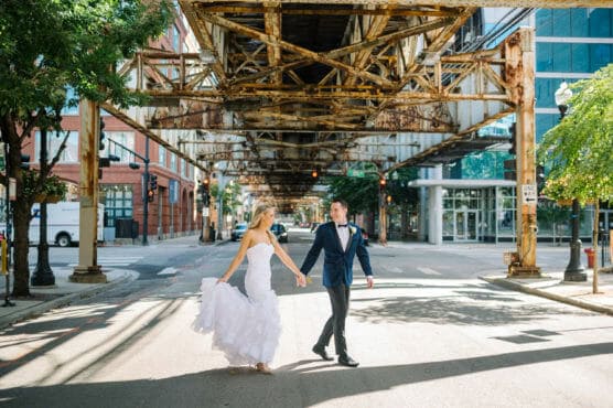 A bride in a white gown and a groom in a dark suit walk hand in hand across a sunlit city street under an elevated train track, surrounded by buildings and trees. by Justin Salem Meyer
