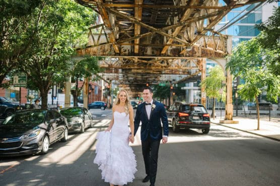A bride in a white dress and a groom in a dark suit walk hand-in-hand down a city street under a rusty elevated train track, surrounded by parked cars and green trees. by Justin Salem Meyer