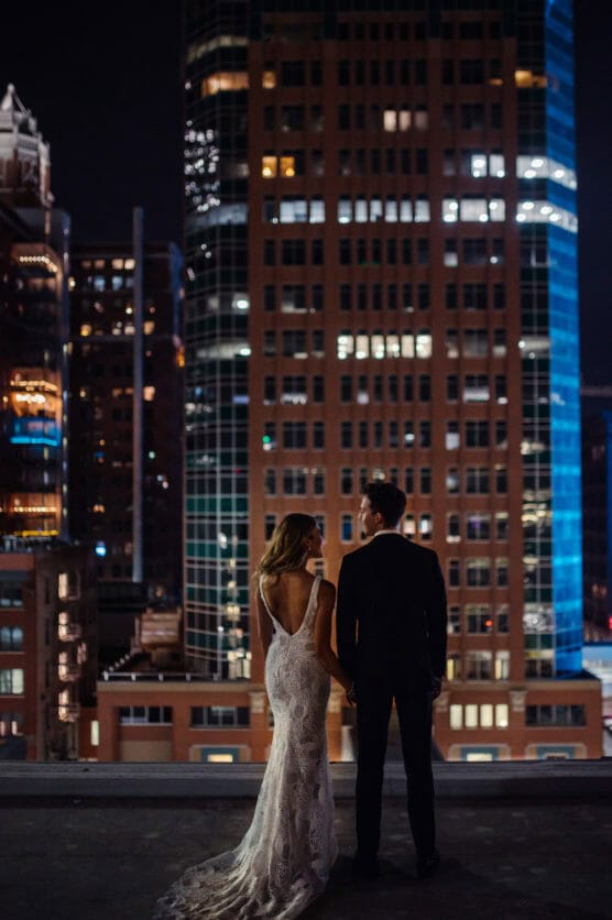 A bride in a lace gown and a groom in a suit stand side by side, facing a city skyline illuminated by lights at night from a rooftop. by Justin Salem Meyer