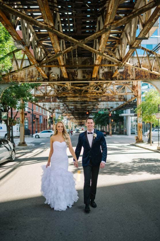 A bride in a white dress and a groom in a dark suit hold hands and walk down a sunlit city street beneath an elevated train track. by Justin Salem Meyer