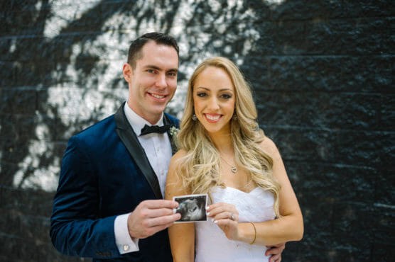 A smiling couple dressed in formal attire stands together outdoors, holding up an ultrasound photo, suggesting a pregnancy announcement. The background features a dark brick wall with dappled sunlight. by Justin Salem Meyer