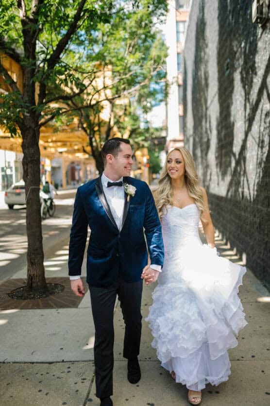 A bride in a white dress and a groom in a blue velvet jacket walk hand-in-hand on a sunny city sidewalk, smiling at each other under leafy trees. by Justin Salem Meyer