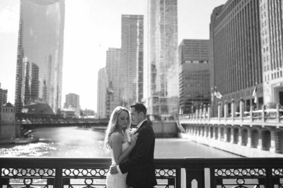 A bride and groom stand close together on a city bridge, surrounded by tall buildings and a river below, with sunlight streaming through, creating a romantic atmosphere. by Justin Salem Meyer