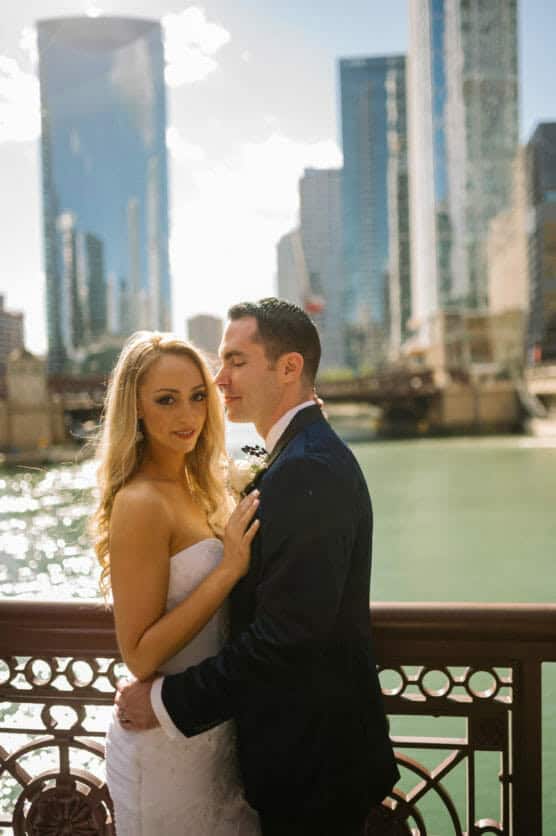 A bride and groom embrace on a city bridge, with tall modern buildings and a river in the background under a bright, sunny sky. by Justin Salem Meyer
