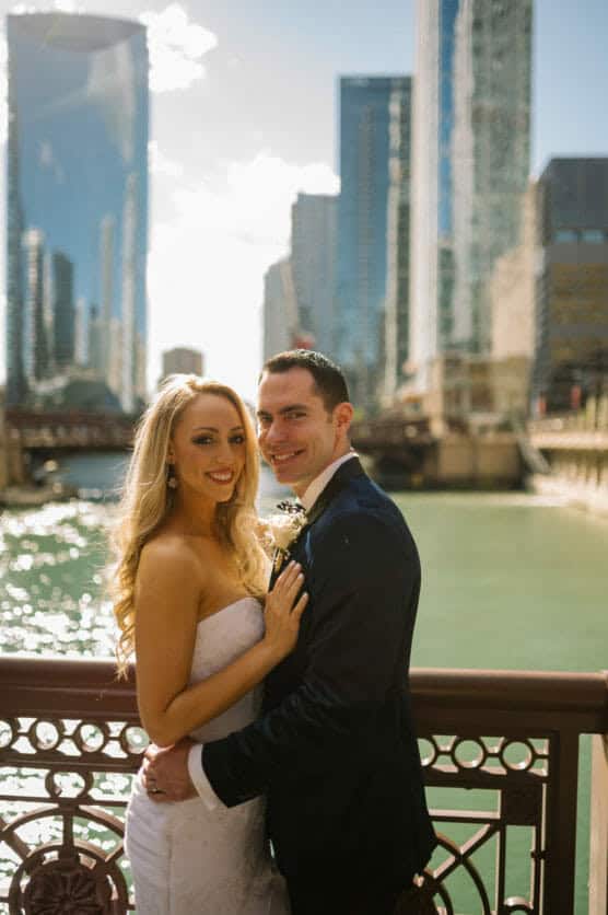 A bride and groom smile and embrace on a sunlit city bridge over a river, with tall skyscrapers and a watery backdrop. The scene suggests a joyful wedding day in an urban setting. by Justin Salem Meyer