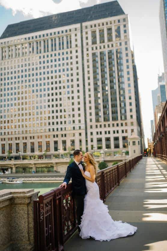 A bride and groom stand close together on a city bridge, surrounded by tall office buildings and a river below, sharing a romantic moment on their wedding day. by Justin Salem Meyer