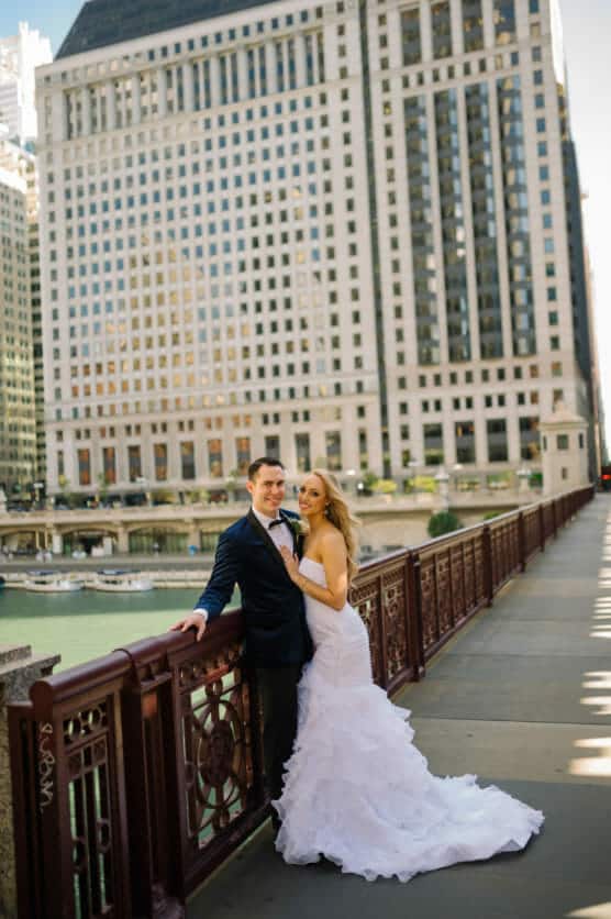 A bride in a white wedding dress and a groom in a dark suit smile and pose together on a city bridge with tall buildings in the background on a sunny day. by Justin Salem Meyer