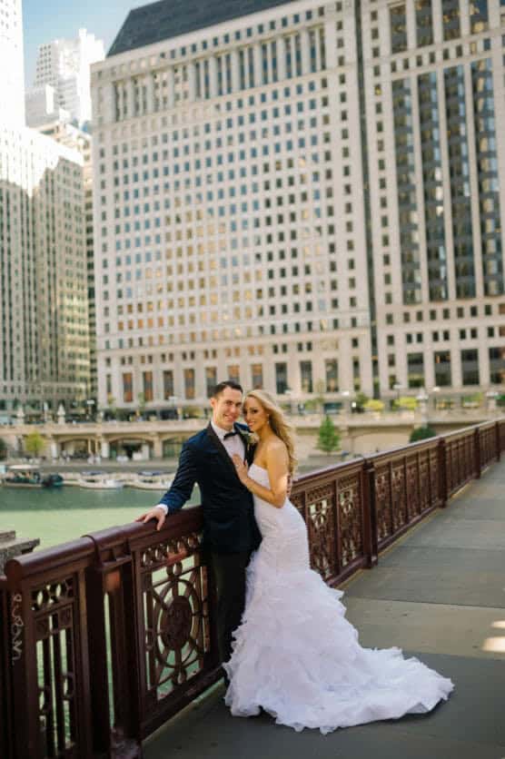 A bride in a white dress and a groom in a dark suit stand close together on a city bridge, with tall buildings and a river in the background on a sunny day. by Justin Salem Meyer