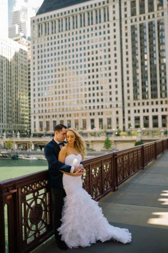 A bride in a strapless white gown and a groom in a dark suit embrace on a city bridge, with tall buildings and water in the background on a sunny day. by Justin Salem Meyer