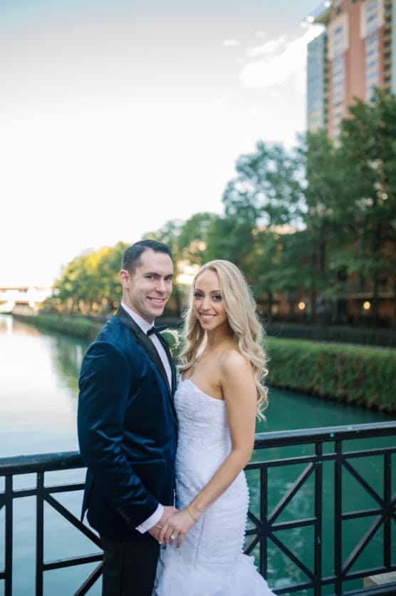 A smiling couple dressed in formal attire pose by a riverside railing, with trees and tall buildings in the background under a clear sky. by Justin Salem Meyer