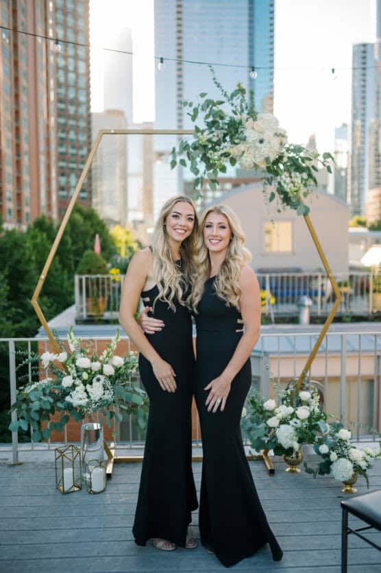 Two women in matching black dresses stand side by side, smiling on a rooftop decorated with white flowers and greenery. Tall city buildings and a gold geometric arch frame the urban background. by Justin Salem Meyer