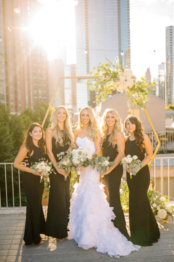 A bride in a white ruffled gown stands with four bridesmaids in black dresses, all holding bouquets, smiling outdoors on a sunny city rooftop with tall buildings in the background. by Justin Salem Meyer