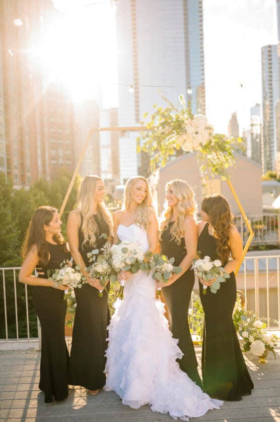 A bride in a white gown stands outdoors with four bridesmaids in black dresses, all holding bouquets and smiling, with sunlight, greenery, and city buildings in the background. by Justin Salem Meyer
