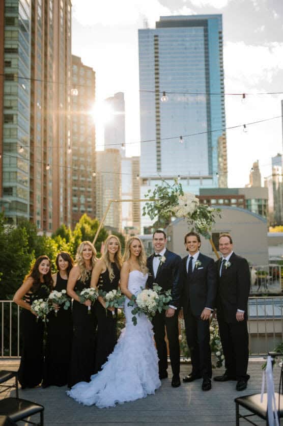 A wedding party poses on a rooftop at sunset, with the bride in a white gown, bridesmaids in black dresses, and groomsmen in black suits, against a city skyline backdrop with tall buildings. by Justin Salem Meyer