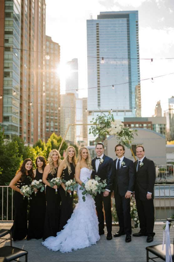 A bride and groom stand together outdoors with three bridesmaids in black dresses and three groomsmen in black suits, posing in front of a city skyline at sunset. by Justin Salem Meyer