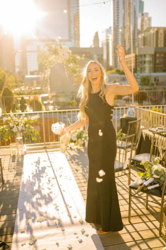 A joyful woman in a black gown tosses flower petals on a sunlit rooftop wedding aisle, laughing with arm raised. City skyline, string lights, and white floral arrangements visible in the background. by Justin Salem Meyer