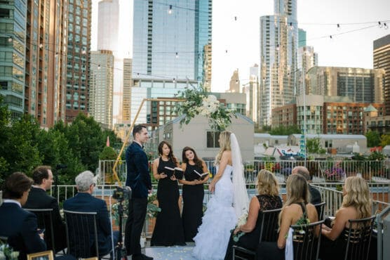 A bride and groom stand facing each other during an outdoor rooftop wedding ceremony in a city, with tall skyscrapers in the background and guests seated watching the event. by Justin Salem Meyer