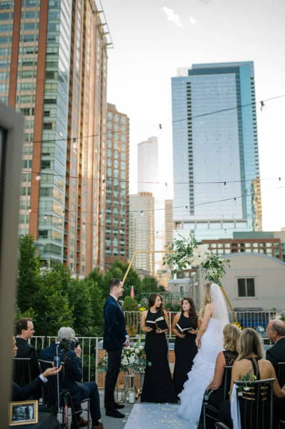 A couple stands at an outdoor rooftop wedding ceremony surrounded by guests. Tall city buildings and string lights form the backdrop. The bride wears a white dress; the groom and bridesmaids wear black attire. by Justin Salem Meyer