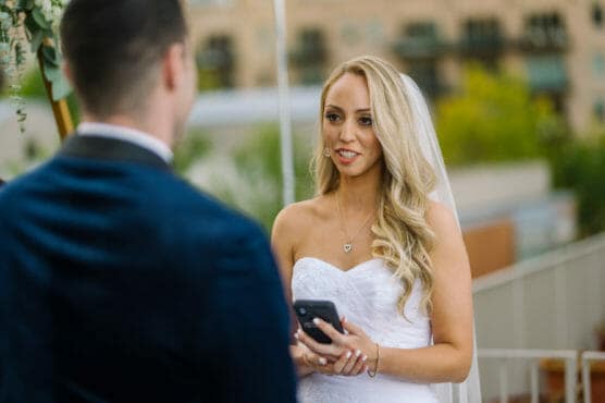 A bride in a white dress and veil holds a phone and smiles while speaking to a groom in a dark suit during an outdoor wedding ceremony. by Justin Salem Meyer