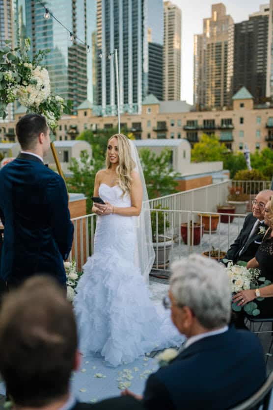 A bride in a white strapless gown and veil stands smiling, holding a note, facing her groom during an outdoor wedding ceremony on a rooftop with city skyscrapers in the background. Guests are seated, watching. by Justin Salem Meyer