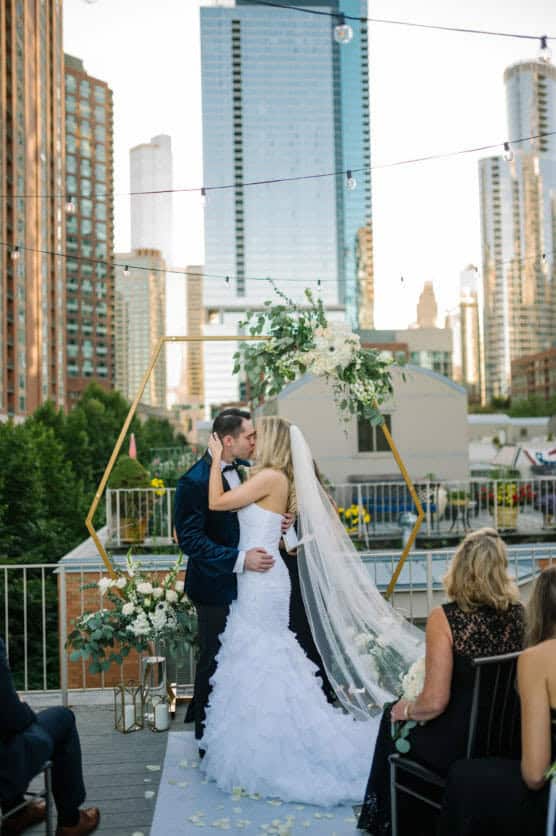 A bride and groom share a kiss during their outdoor rooftop wedding ceremony, surrounded by tall city buildings and floral decorations, as guests watch from their seats. by Justin Salem Meyer