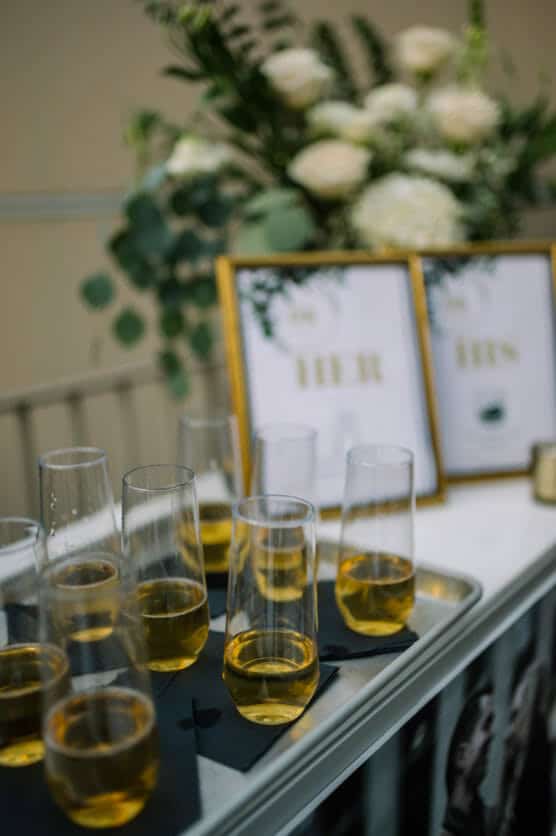 A tray of champagne flutes filled with a golden drink sits on a table. In the background, white flowers, greenery, and two framed signs labeled HER and HIS are visible. by Justin Salem Meyer