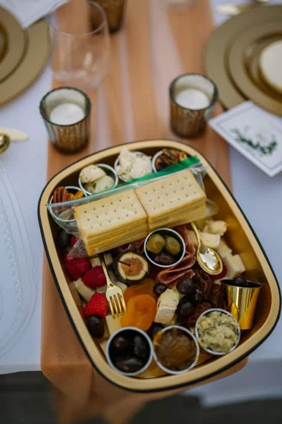 A tray with assorted crackers, cheeses, nuts, dried fruits, fresh berries, olives, and dips, arranged neatly on a table set with candles, plates, gold cutlery, and elegant tableware. by Justin Salem Meyer