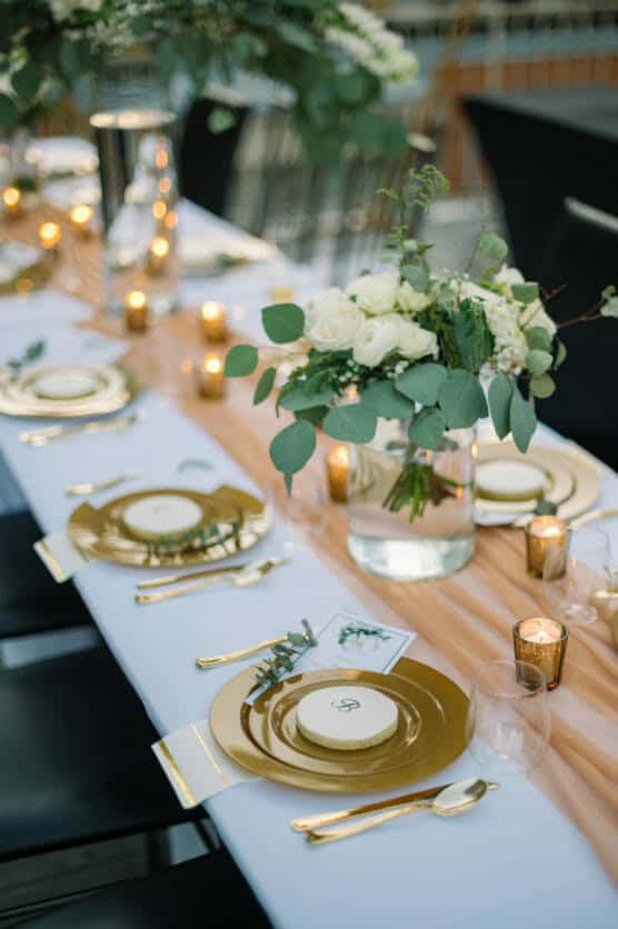 A beautifully set table with gold plates and cutlery, white napkins, small floral arrangements with white roses and eucalyptus, and lit votive candles, creating an elegant and inviting atmosphere. by Justin Salem Meyer