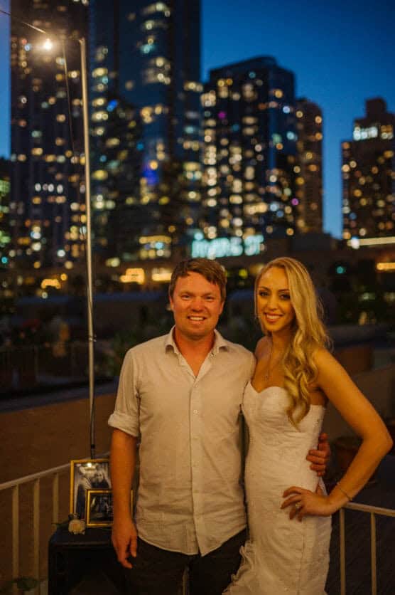 A man and a woman in formal attire pose and smile on an outdoor balcony at night, with a city skyline and tall buildings lit up in the background. by Justin Salem Meyer