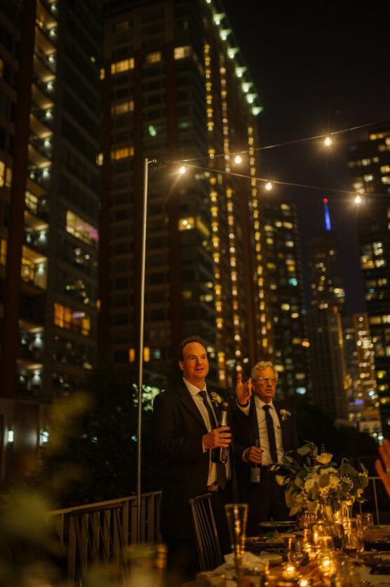 Two men in suits stand on a rooftop at night, one holding a drink and the other gesturing, surrounded by string lights. Tall, illuminated buildings create a city backdrop, while a decorated table is visible in front of them. by Justin Salem Meyer