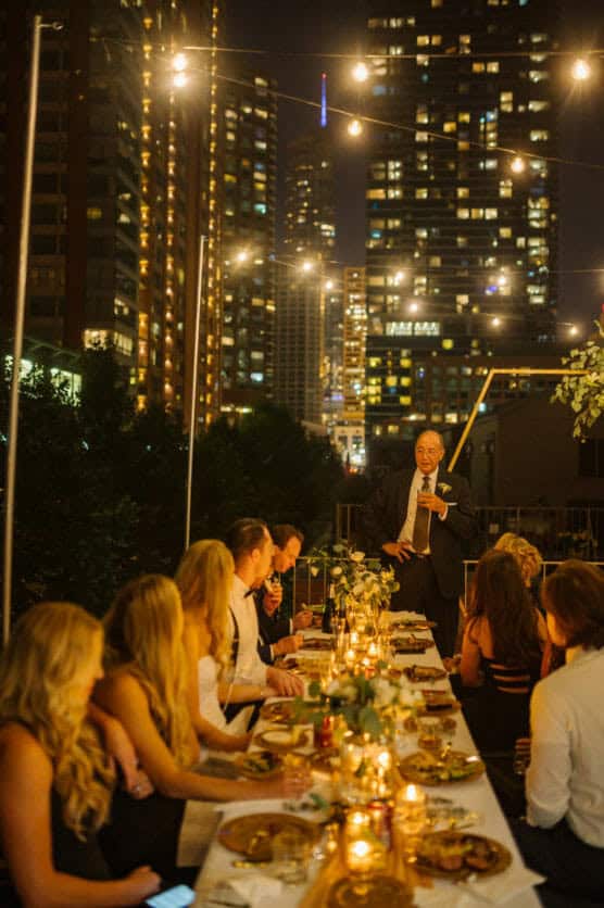 A group of people dine at a long, decorated outdoor table under string lights at night, with a man standing and speaking, and tall city buildings illuminated in the background. by Justin Salem Meyer