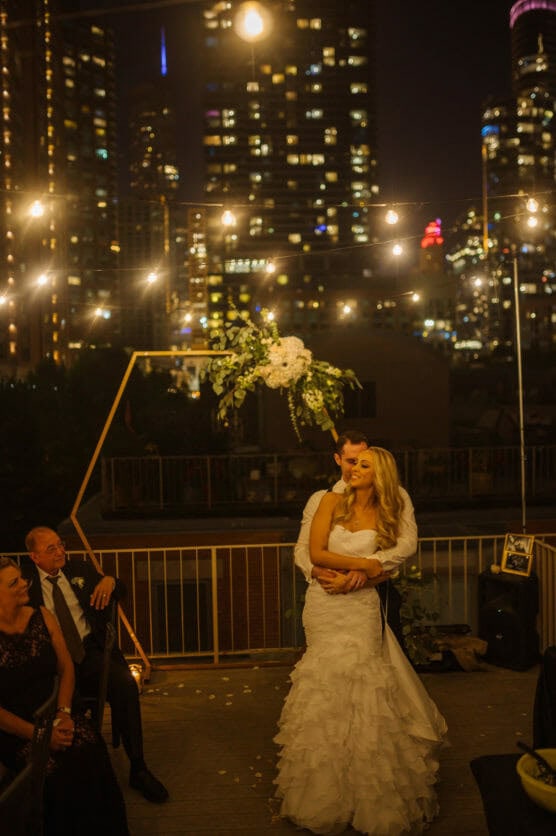 A bride and groom share a slow dance at an outdoor rooftop wedding at night, surrounded by string lights and tall city buildings, with guests seated nearby watching them. by Justin Salem Meyer