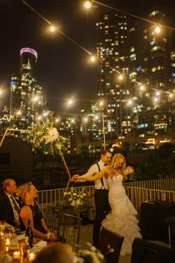 A bride and groom dance under string lights at a nighttime outdoor wedding reception, with guests seated at tables and city skyscrapers glowing in the background. by Justin Salem Meyer