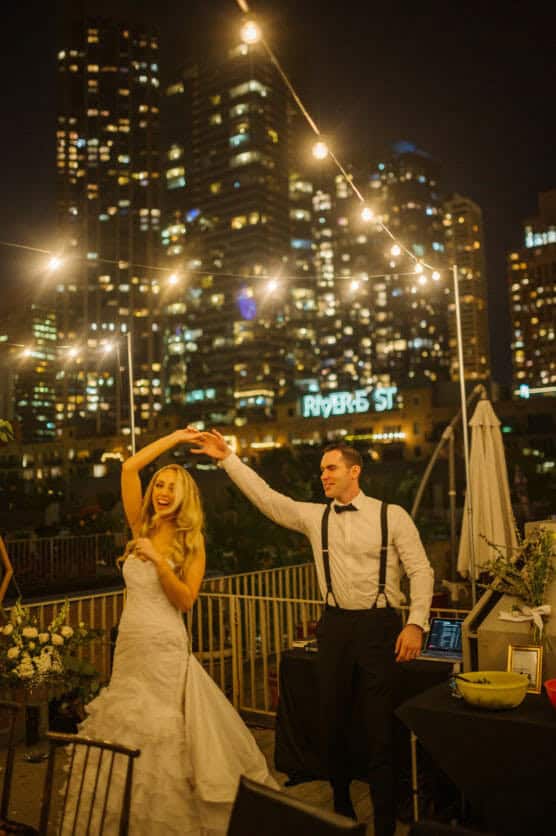 A bride in a white gown and a groom in suspenders dance joyfully outdoors at night, under string lights, with city skyscrapers and a RIVER ST sign glowing in the background. by Justin Salem Meyer