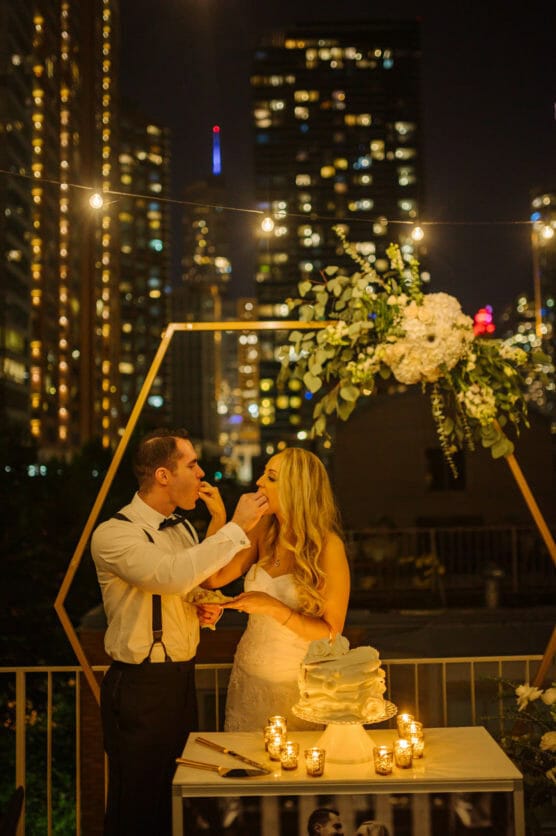 A bride and groom feed each other cake at night on a city rooftop decorated with flowers and string lights, with tall buildings illuminated in the background. by Justin Salem Meyer