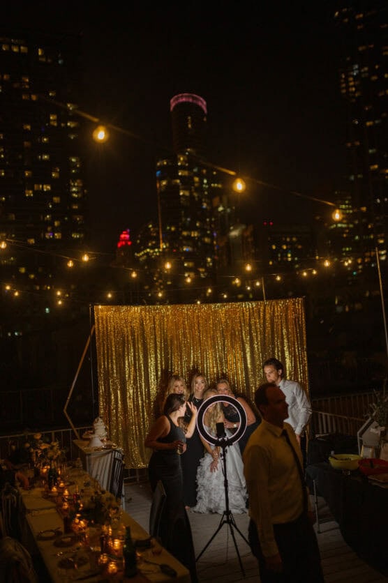 Four people pose for a photo in front of a gold sequin backdrop at a nighttime rooftop event, with city lights and string lights illuminating the scene. A ring light and camera are set up in front of them. by Justin Salem Meyer