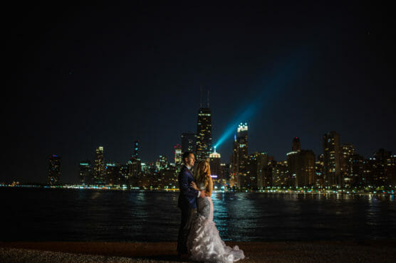 A couple in formal attire embraces at night by the water, with brightly lit city skyscrapers and reflections in the background. by Justin Salem Meyer