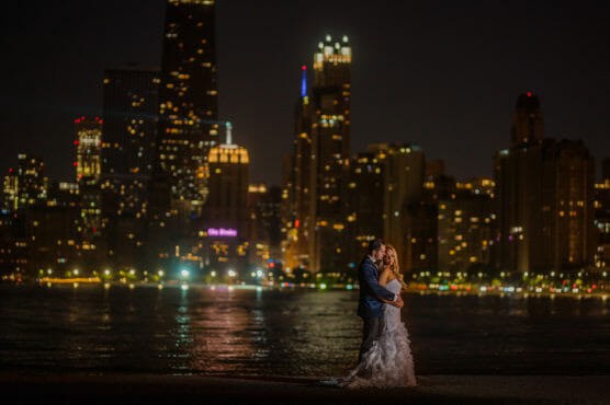 A bride and groom embrace by the water at night, with a city skyline illuminated by lights in the background. by Justin Salem Meyer