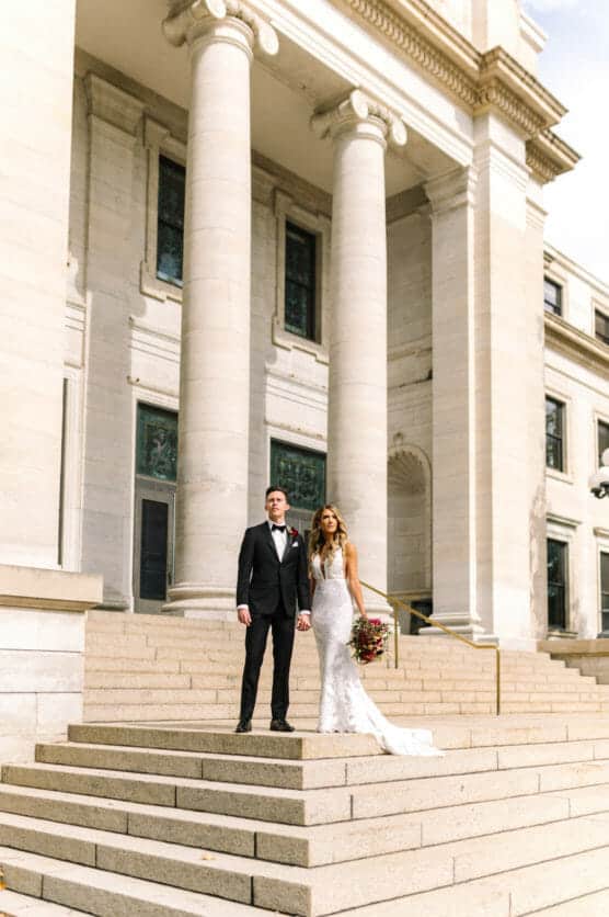 A bride in a white gown and a groom in a black tuxedo stand together on the steps of a grand building with tall columns, holding hands. The bride holds a bouquet of flowers. by Justin Salem Meyer