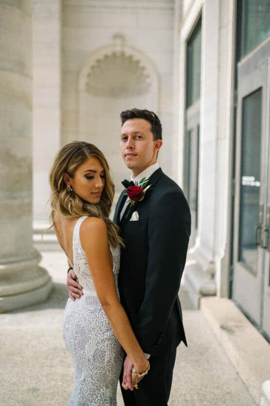 A bride in a lace dress and a groom in a black tuxedo stand closely together, holding hands, with neutral expressions. They are in an elegant, light-filled building with stone columns and arched details. by Justin Salem Meyer