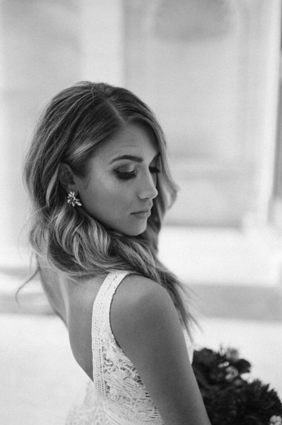 A woman in a lace wedding dress with wavy hair looks down over her shoulder, holding a bouquet. The photo is black and white and set against a softly blurred background. by Justin Salem Meyer